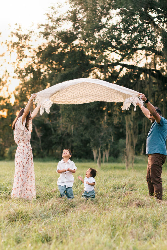 family playing parachute with a blanket in a field in jacksonville with two little boys standing underneath the blanket laughing