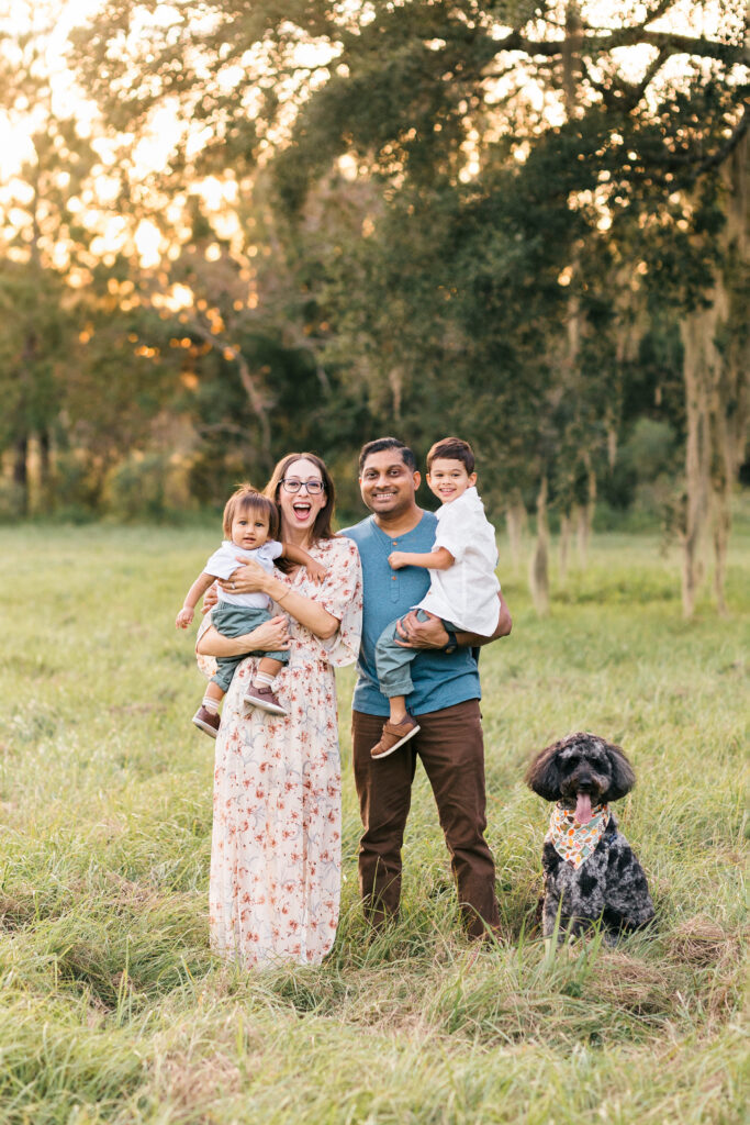 a mom and dad stand in a field in Jacksonville, holding their two little boys and laughing; their dog is sitting beside them
