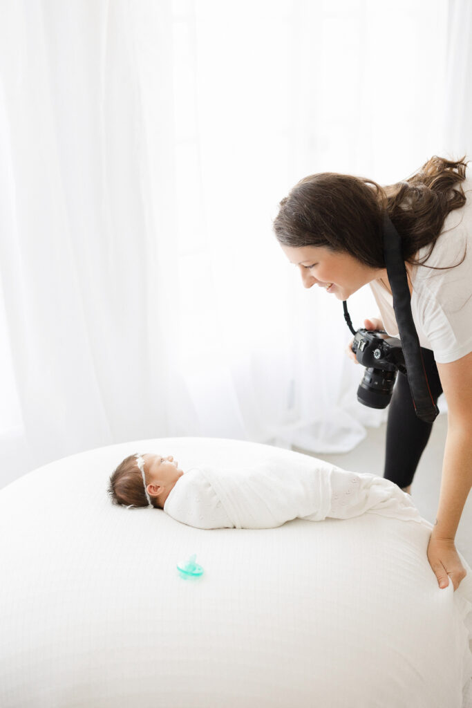 a newborn photographer smiles and holds a camera as she leans over a baby swaddled in a white blanket