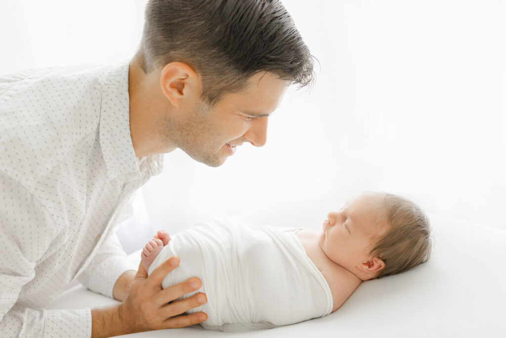 a dad leans over and stares lovingly at his swaddled, newborn baby girl; she is on a white cushion and the background is pure white light