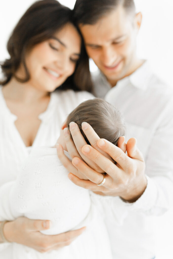 close up of mom and dad's hands holding their newborn baby while they smile in the background
