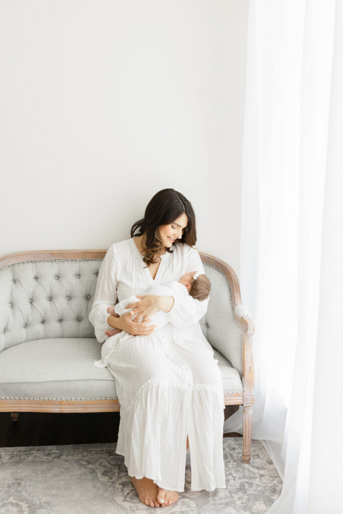 a mom in a long white dress sits on a gray couch smiling and holding her new daughter in her arms