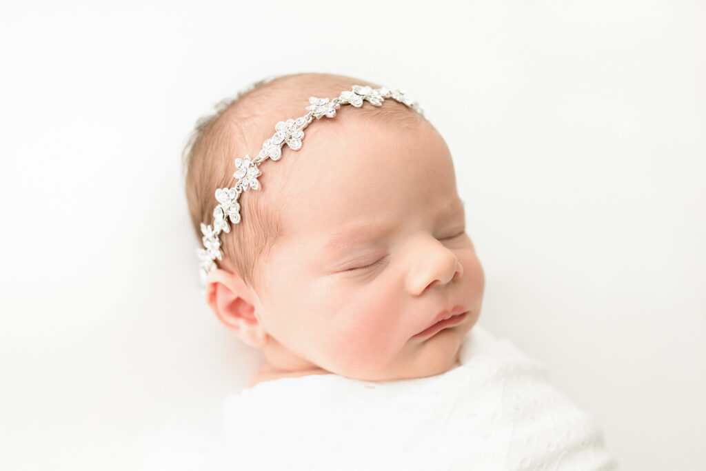 a newborn baby girl lays sleeping on a white bed; she is wearing a dainty silver and crystal headband