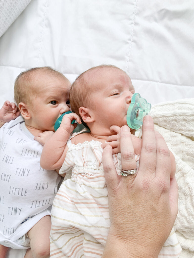 boy/girl newborn twins lay on a white blanket; they have blue pacifiers in their mouths